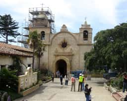 Front of the Carmel Mission Basilica with its two bell towers, one under scaffolding, and visitors in the forecourt