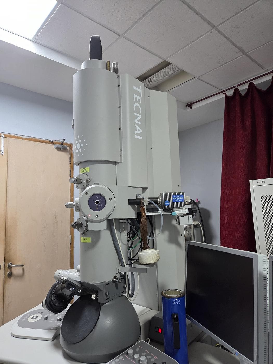 A tall grey transmission electron microscope column with a viewing chamber, control panels and a monitor in a research laboratory