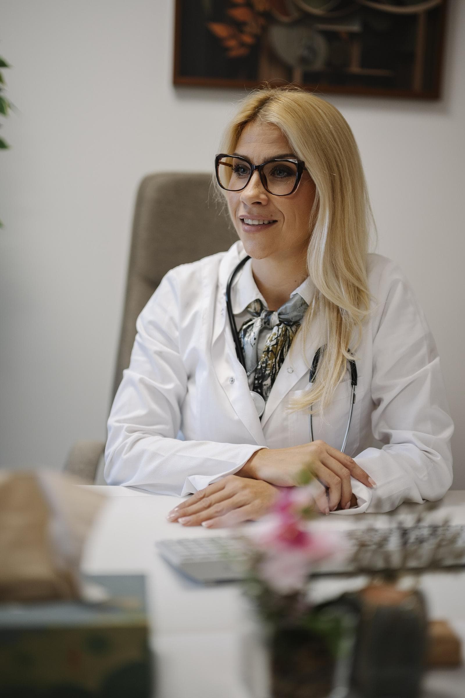 Clinician in a white coat and glasses seated at a consultation desk with flowers in the foreground