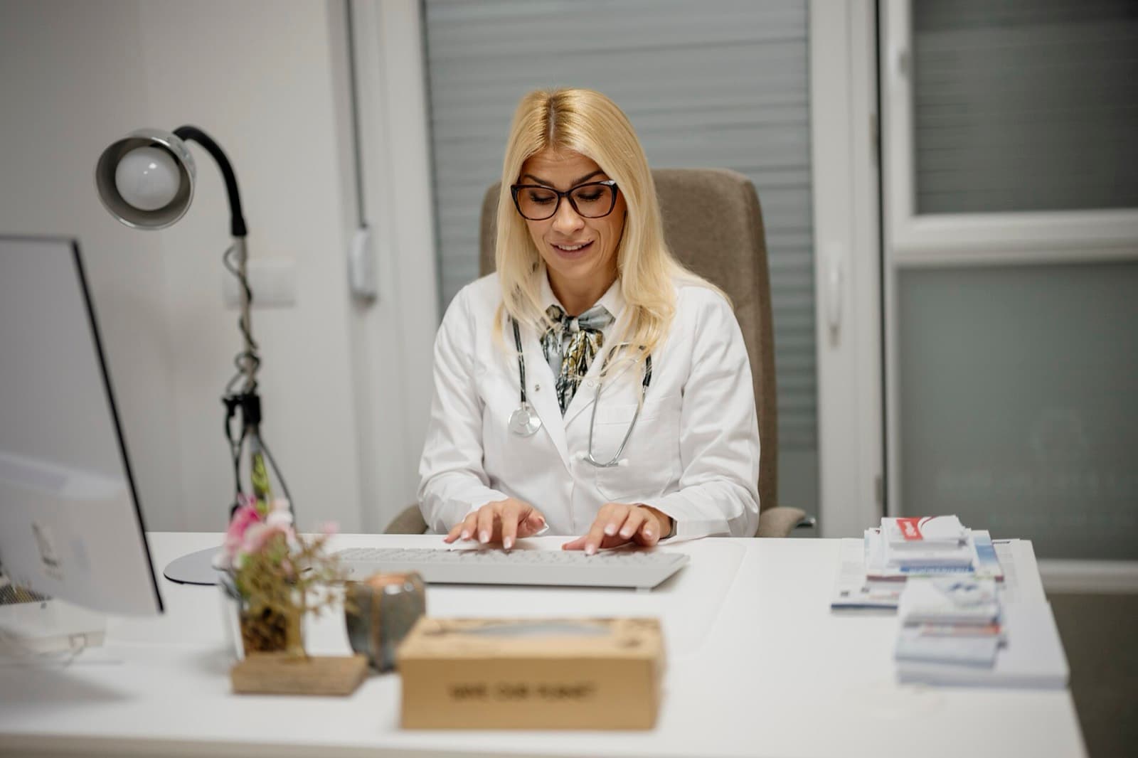 Clinician in a white coat typing at a computer behind a white desk in a bright consultation office