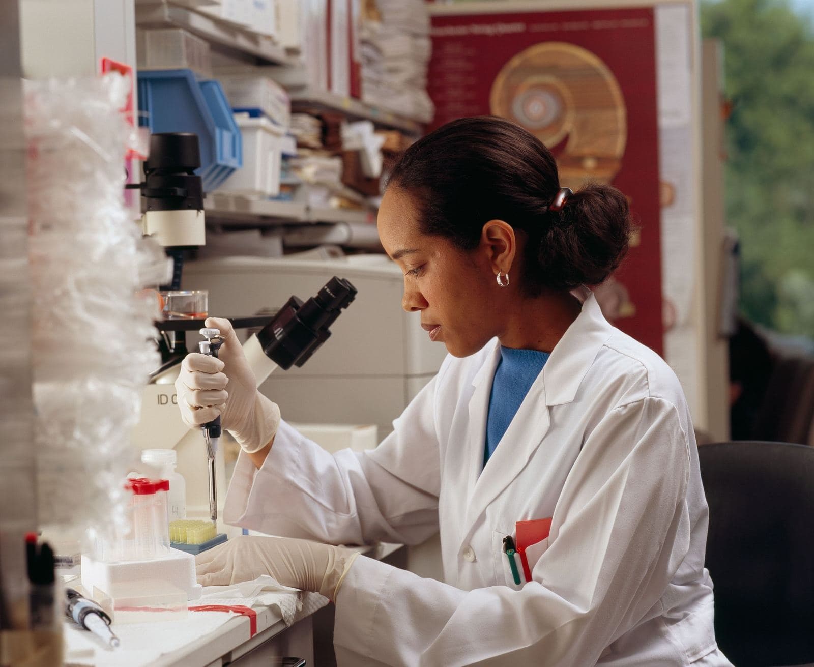 Researcher in a white lab coat and gloves using a pipette at a laboratory bench beside a microscope and sample racks