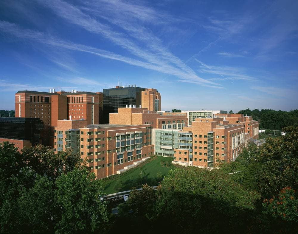 Aerial photograph of the Mark O. Hatfield Clinical Research Center, Building 10, on the NIH campus in Bethesda, Maryland: red brick wings around a central tower, a lawn in front and trees in the foreground under a blue sky