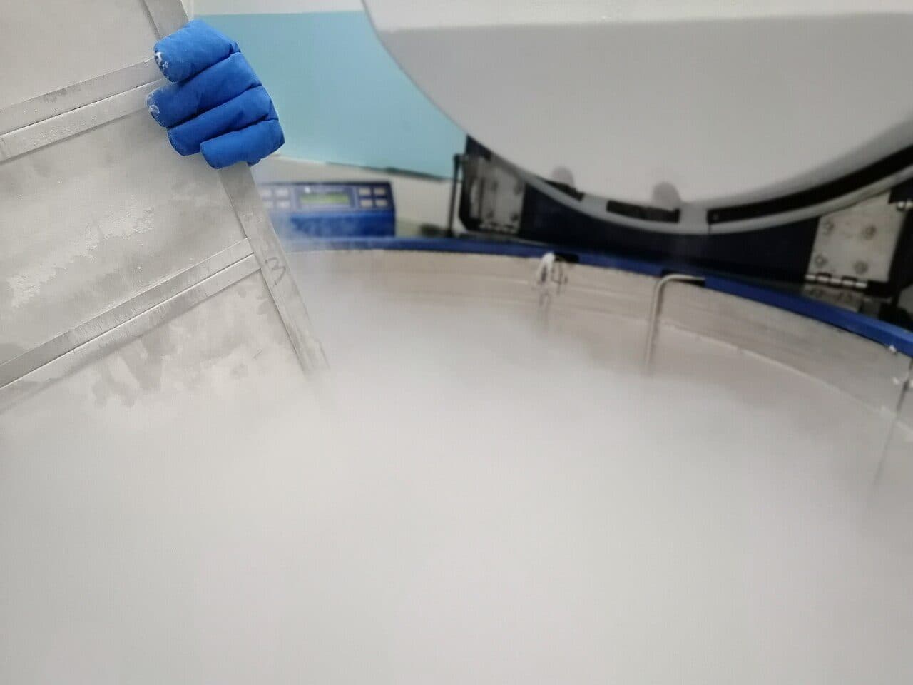 A gloved hand in an insulated cryogenic glove lifts a metal storage rack from a liquid nitrogen freezer, with vapor rising from the open vessel in a hospital cell processing room.