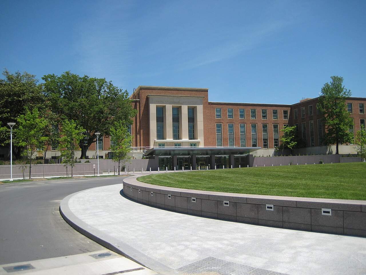 Brick and limestone building with a columned central entrance and glass canopy, seen across a curved granite-edged lawn under a clear blue sky at the FDA White Oak campus in Silver Spring, Maryland.